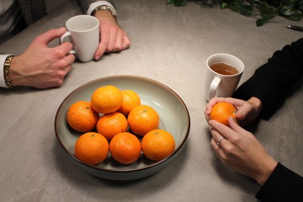 Gesprek tussen twee mensen, waarbij een fruitmand met mandarijnen en twee kopjes thee op tafel staan.